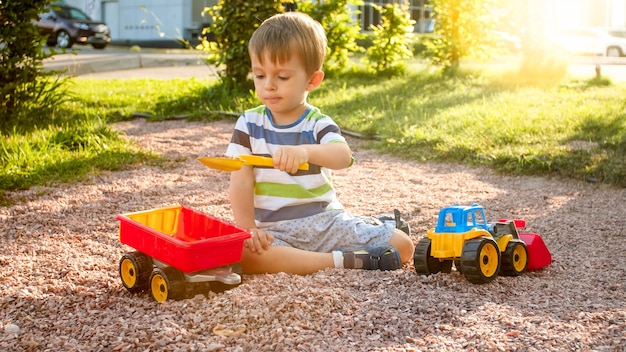 Nahaufnahmeporträt eines glücklich lächelnden 3 Jahre alten Kinderjungen, der Sand auf dem Spielplatz mit Spielzeugplastik-LKW oder Bagger gräbt