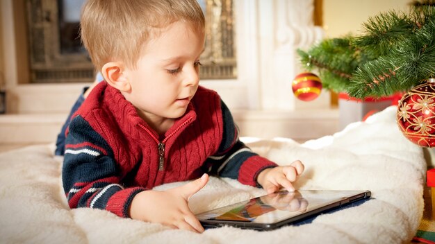 Nahaufnahmeporträt eines glücklich lächelnden kleinen Jungen mit digitalem Tablet-Computer beim Liegen auf dem Boden unter dem Weihnachtsbaum im Wohnzimmer