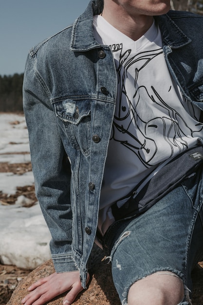 Nahaufnahmeporträt eines Hipsters, der im Winter auf dem Stein sitzt und Jeans und ein T-Shirt trägt.