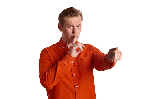 Nahaufnahmeporträt eines jungen athletischen rothaarigen Kerls in einem stylischen orangefarbenen Hemd, der jemanden ansieht und ein leises Zeichen zeigt, während er isoliert auf weißem Studiohintergrund posiert. Menschliche Gesichtsausdrücke. Aufrichtig