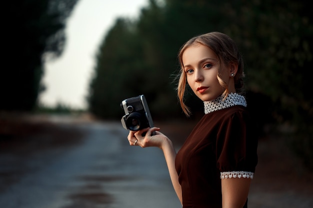 Nahaufnahmeporträt eines jungen schönen Mädchens in einem braunen Kleid im Retro-Stil mit einer Vintage-Kamera in ihren Händen auf einer verlassenen Straße.