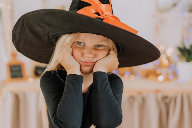 Nahaufnahmeporträt eines kleinen blonden Mädchens mit blauen Augen in einem schwarzen Hut in den Halloween-Dekorationen