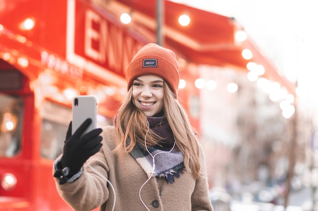 Nahaufnahmeporträt eines lächelnden Mädchens in der warmen Kleidung nimmt selfie auf einem Smartphone mit einem hellen roten Stadthintergrund