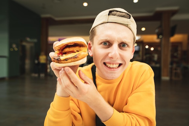 Nahaufnahmeporträt eines lustigen jungen Mannes, der einen Burger in seiner Hand hält und die Kamera untersucht
