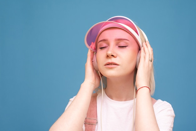 Nahaufnahmeporträt eines Mädchens in einem weißen T-Shirt und einer rosa Kappe, hört Musik in ihren Kopfhörern mit ihren geschlossenen Augen auf Blau