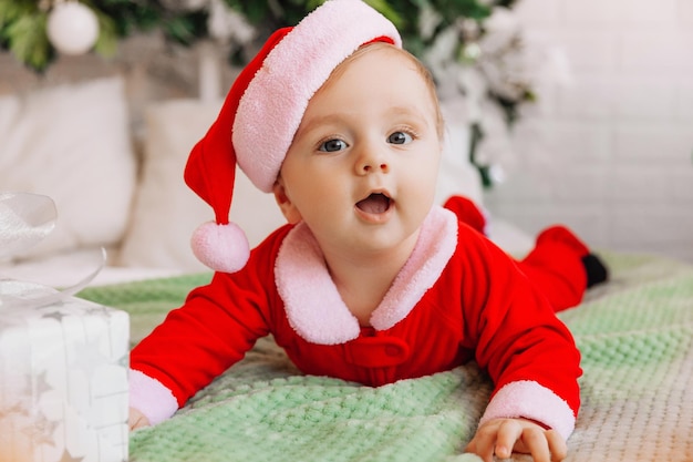 Nahaufnahmeporträt eines Neugeborenen. Kaukasisches Baby 6 Monate in einem Weihnachtsmannanzug, der auf einer gestrickten Kuscheldecke auf dem Bett liegt. Weihnachten 2022