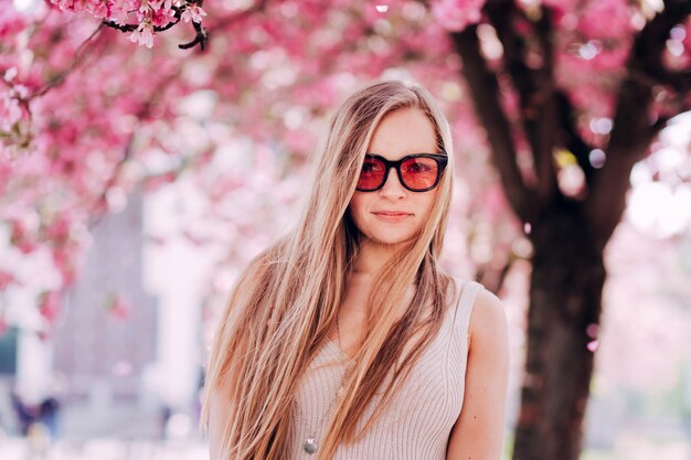 Nahaufnahmeporträt eines reizenden Mädchens in einem Park mit blühenden japanischen Sakura-Bäumen. Romantische junge Blondine in einem Kleid und in der rosa Brille, die auf einem Hintergrund der Frühlingsblumenbäume aufwirft. Kirschblüten.