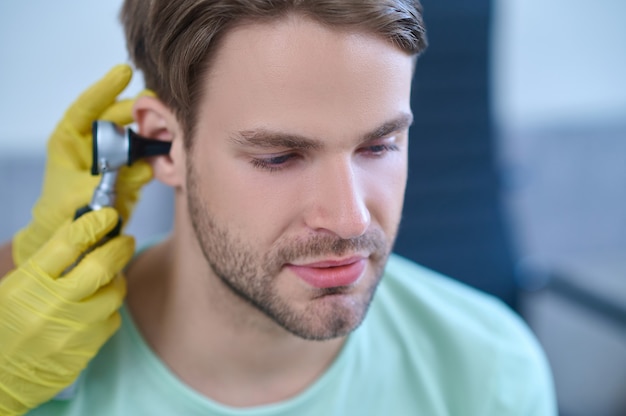 Foto nahaufnahmeporträt eines ruhigen, dunkelhaarigen, bärtigen, attraktiven jungen mannes, der sich einer ärztlichen untersuchung unterzieht, die von einer erfahrenen hno-ärztin durchgeführt wird