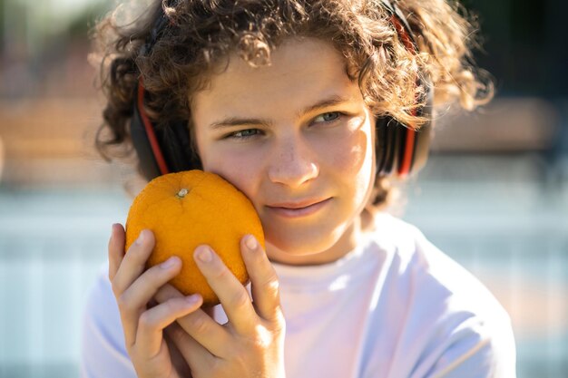 Nahaufnahmeporträt eines süßen, verträumten Teenagers in kabellosen Kopfhörern, der die Orange gegen sein Gesicht drückt