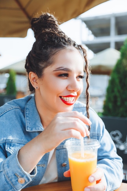 Nahaufnahmeporträt im Freien eines netten modischen Mädchens, das Spaß hat, Orangensaft in einem Café im Freien zu trinken, eine Frau mit Dreadlocks und rotem Lippenstift