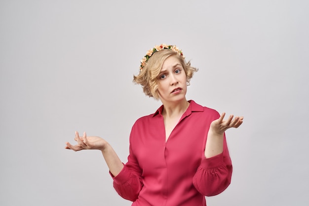 Nahaufnahmeporträt im Studio auf weißem Hintergrund verwirrte gestörtes junges Mädchen in einem roten Kleid, das Hände vor ihr in Verwirrung, Zweifel, Protest trennt. Blondes Haar und Blumenkranz auf ihrem Kopf