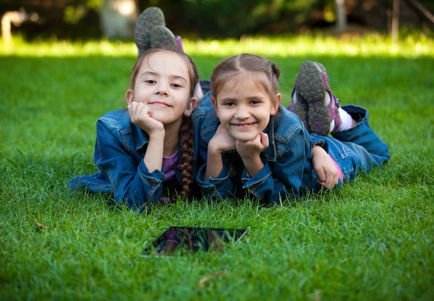 Nahaufnahmeporträt von zwei kleinen Mädchen, die mit Tablet auf Gras liegen