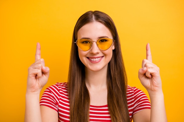 Nahaufnahmeporträtfoto des süßen, reizenden jungen Promotermädchens, das lächelt und mit den Fingern auf leeren Raum zeigt, schlagen vor, tragen Sie Sonnenspezifikationen, gestreiftes weißes rotes Hemd, leuchtend gelber Farbhintergrund