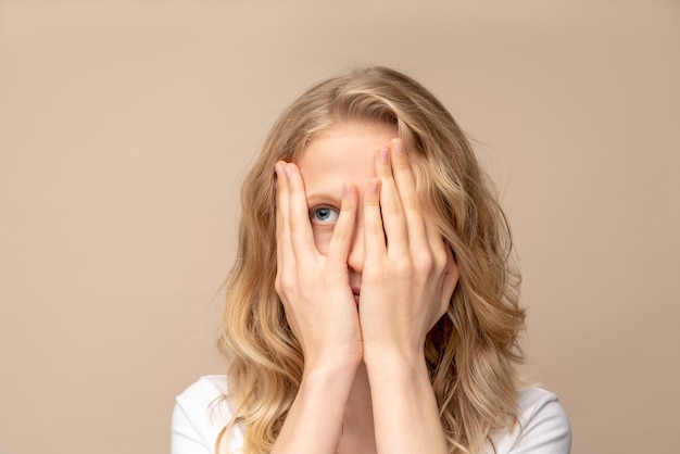 Nahaufnahmeporträtfrau, die Hand auf Gesicht bedeckt, guckend mit einem Auge. Auf beige Wand isoliert.