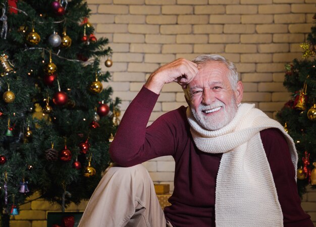 Nahaufnahmeportrait des lächelnden hübschen kaukasischen Älteren Mannes im roten Pullover und im Schal, der vor geschmücktem Weihnachtsbaum sitzt. Winterurlaub entspannen.