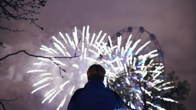 Nahaufnahmesilhouette eines Mannes, der Feuerwerke beobachtet und fotografiert, die auf der Smartphone-Kamera im Freien explodieren