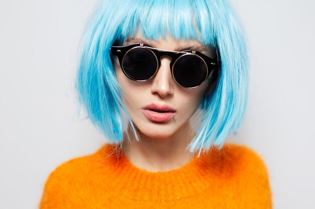 Nahaufnahmestudioporträt des modischen hübschen Mädchens mit dem blauen Haar, das runde Sonnenbrille und das orange Hemd auf weißem Hintergrund trägt.