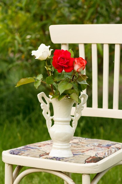 Nahaufnahmevase mit Rosenblüten auf einem weißen rustikalen Stuhl im Garten mit natürlichem Hintergrund.