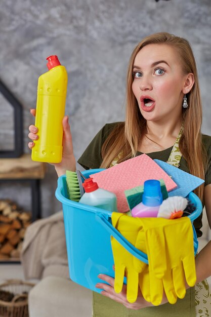 Nahaufnahmewaschmittel zur Reinigung emotionaler Frauen- oder Hausfrauenhände mit vielen Haushaltsgegenständen in blauem Eimer im Wohnzimmer. Hausaufgaben. Platz für Text auf der Flasche