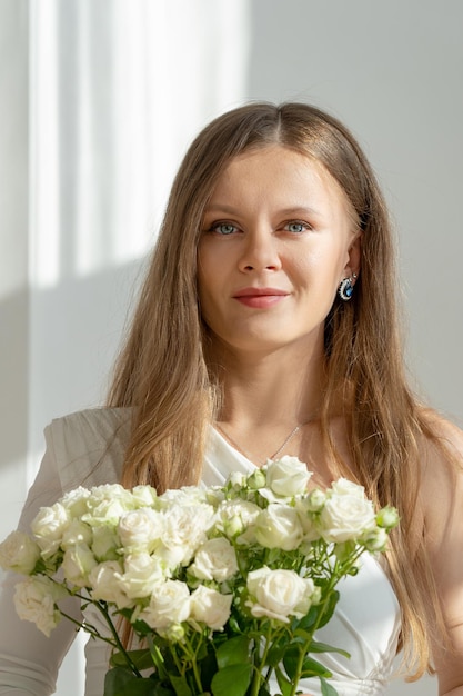 Nahe Porträt einer eleganten Braut mit blonden Haaren mit Blumen in weißem Kleid, die in die Kamera schaut