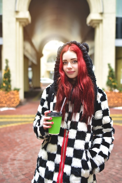 Nahe Porträt eines jugendlichen Mädchens mit roten Haaren in der warmen Kleidung, die draußen an einem kalten Tag mit einer Tasse Kaffee in ihren Händen steht und in die Kamera schaut.