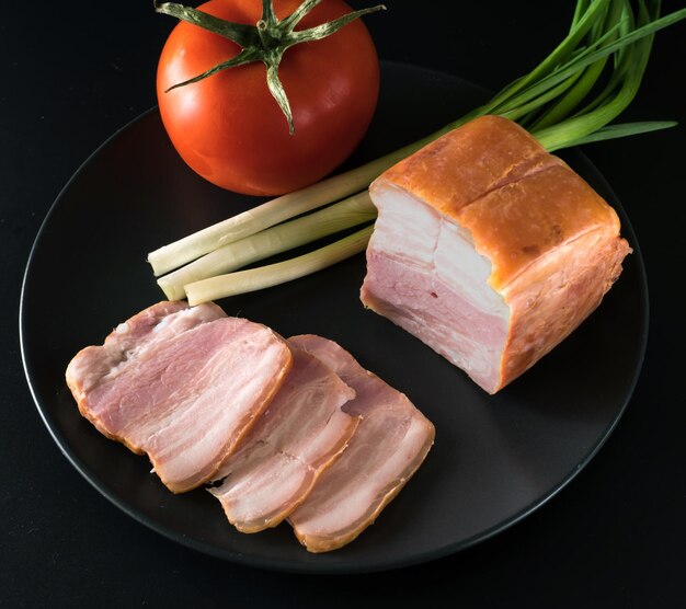 Nahrungsmittel geschnittenes Speck, Tomaten, grüne Zwiebeln auf einem schwarzen Teller auf schwarzem Hintergrund