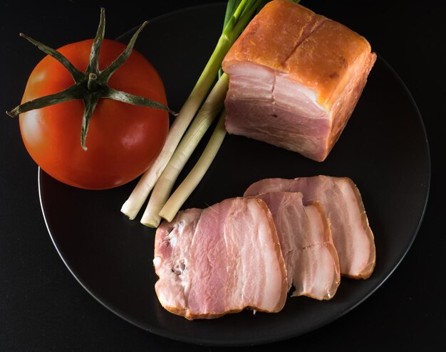 Nahrungsmittel geschnittenes Speck, Tomaten, grüne Zwiebeln auf einem schwarzen Teller auf schwarzem Hintergrund