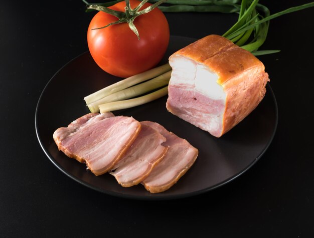 Nahrungsmittel geschnittenes Speck, Tomaten, grüne Zwiebeln auf einem schwarzen Teller auf schwarzem Hintergrund