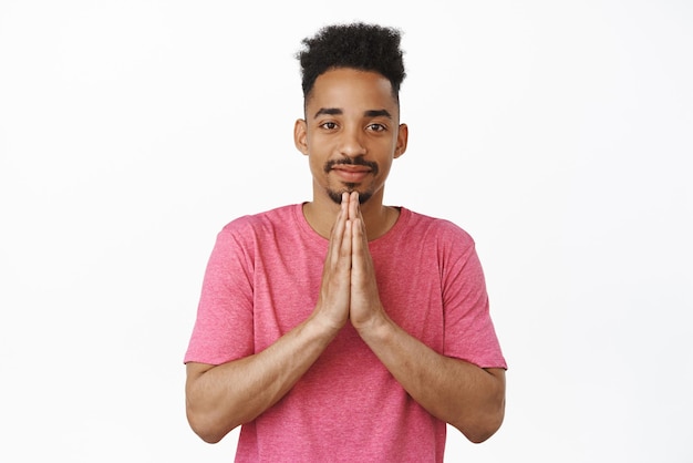 Namaste mi amigo Chico afroamericano sonriente con peinado rizado tomados de la mano en gesto de súplica de oración inclinándose cortésmente de pie en camiseta rosa contra fondo blanco