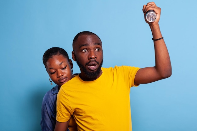 Namorada com medo se escondendo atrás do namorado com lanterna, verificando o problema. Casal apavorado com som de terror, tendo expressão facial perturbada. Pessoas em pé sobre o fundo.