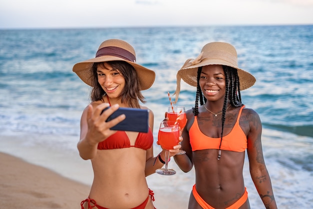 Namoradas contentes com bebidas fazendo selfie na praia