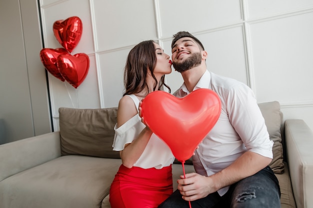 Foto namorado e namorada com balão em forma de coração, sorrindo e sentado no sofá em casa