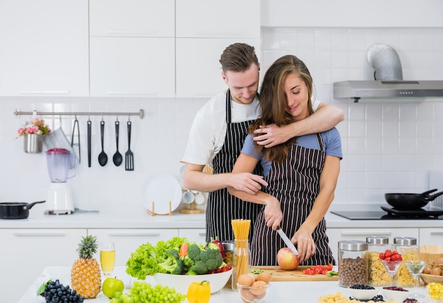 Namorado sorridente, abraça a mulher por trás, vendo-a preparar salada de frutas na cozinha, um homem amoroso, abraça a namorada, cortando frutas frescas