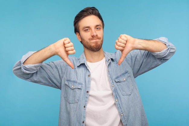 Não gostei, péssimo atendimento. Retrato de homem insatisfeito em camisa jeans trabalhador mostrando os polegares para baixo, expressando desaprovação, criticando e discordando da sugestão. tiro de estúdio interno, fundo azul