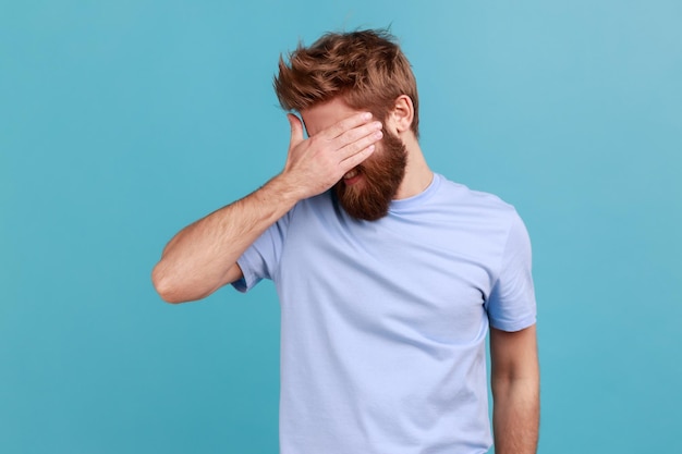 Não quero olhar para este retrato de homem barbudo cobrindo os olhos e virando com desgosto de algo vergonhoso com medo de ver tiro de estúdio interior isolado em fundo azul