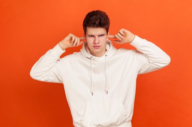 Não quero ouvir Adolescente irritado em moletom elegante com capuz fechando as orelhas com os dedos desobediência ignorando ruído irritante Foto de estúdio interior isolada em fundo laranja