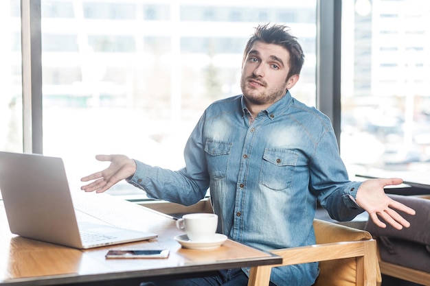 Não tenho certeza! Retrato de confuso bonito barbudo jovem freelancer em camisa jeans azul está sentado no café e trabalhando no laptop com os braços erguidos e rosto perplexo, olhando para a câmera. interior