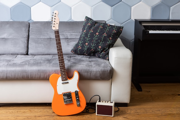 Naranja guitarra eléctrica y amplificador con cable cerca de un sofá de cuero y piano en la sala de estar