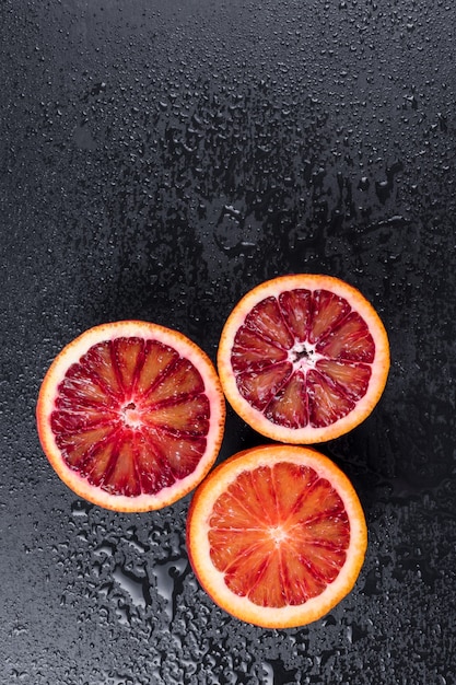 Naranjas ensangrentadas cortadas por la mitad en pizarra negra con gotas de agua, fruta naranja roja siciliana, vista superior, espacio libre para texto.