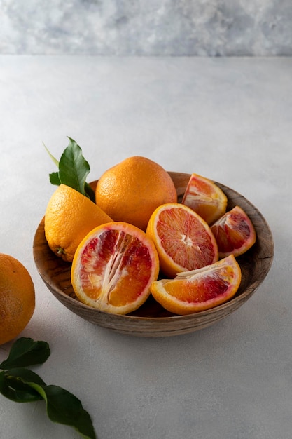 Naranjas sangrientas en tazón de madera, sobre fondo brillante, enteras y rebanadas con hojas de cítricos frescos