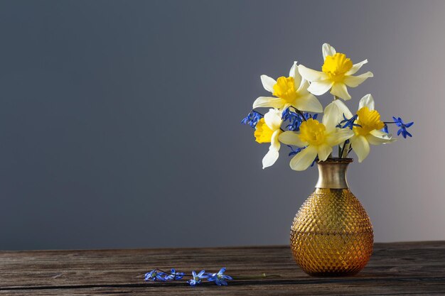 Narciso amarillo y campanillas azules en jarrón sobre mesa de madera sobre fondo oscuro