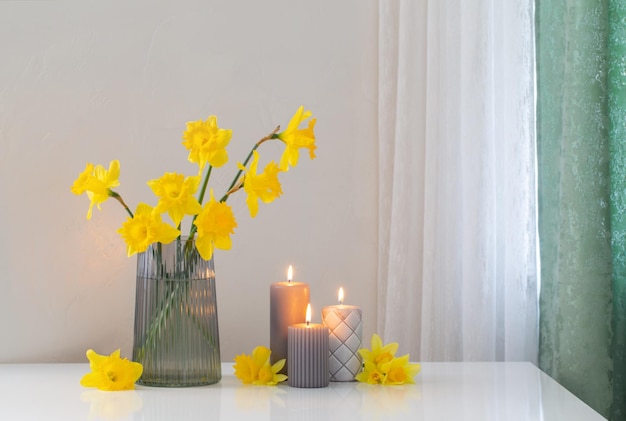 Narcisos amarelos da primavera em vaso de vidro com velas acesas no fundo branco