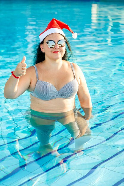 Nas férias, uma mulher com chapéu de Papai Noel, óculos escuros e maiô na piscina mostra sua classe com a mão. Foto vertical