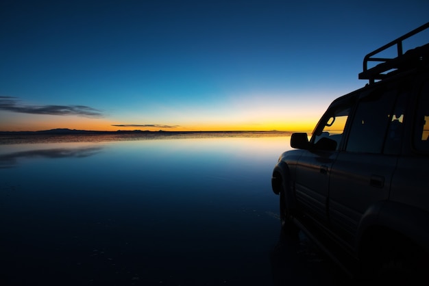 Nascer do sol no Salar de Uyuni na Bolívia coberto de água, carro no deserto salgado e reflexos do céu