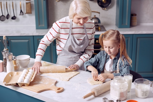 Naschkatze. Entzückendes kleines Mädchen, das ihrer Großmutter hilft, Kekse zu machen, verschiedene Figuren herauszuschneiden, während die Frau etwas mehr Teig ausrollt