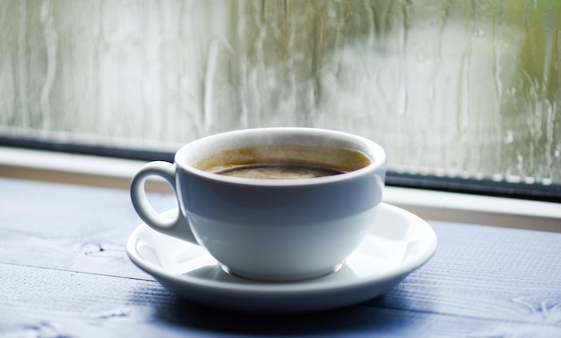 Nasses Glasfenster und Tasse heißen Kaffee. Herbsttrübes Wetter besser mit Koffeingetränk. Kaffee an regnerischen Tagen genießen. Frisch gebrühter Kaffee in weißer Tasse oder Becher auf der Fensterbank. Kaffeezeit an regnerischen Tagen.