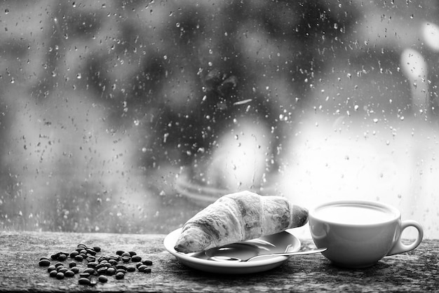 Nasses Glasfenster und Tasse heißes Koffeingetränk Kaffeegetränk mit Croissant-Dessert Kaffeegenuss an regnerischen Tagen Kaffeezeit an regnerischen Tagen Frisch gebrühter Kaffee in weißer Tasse oder Becher auf der Fensterbank