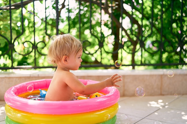 Nasses Kind sitzt in einem kleinen aufblasbaren Pool und fängt Seifenblasen