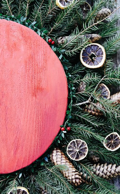 Natal, ano novo, composição vertical do feriado de inverno com superfície de madeira vermelha em galhos de árvore verde do abeto, fatias de laranja secas, cones e laço vermelho.