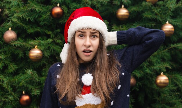 Natal chocado ou surpreso. Feche o retrato de uma mulher com um chapéu de Papai Noel com emoção. No contexto de uma árvore de Natal.